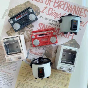May include: A collection of miniature kitchen and electronic appliances including two grey and red boombox radios, two white toaster ovens with glass doors, and two rice cookers in white and light blue with black lids and control panels.