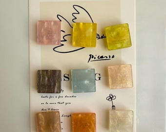 Puede incluir: Nueve imanes decorativos cuadrados en varios colores, incluyendo rosa, amarillo, marrón y naranja. Cada imán tiene una apariencia ligeramente translúcida y presenta el texto "CREATION FRANCE". Los imanes están dispuestos en una tarjeta con un dibujo al estilo Picasso.