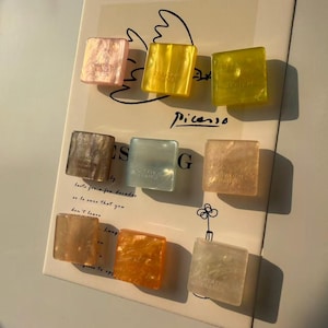 May include: Nine square-shaped hair clips in various colors, including pink, yellow, and orange. Each clip has a marbled effect and the text "CREATION FRANCE". The clips are arranged on a paper with the word "Picasso" and other text.