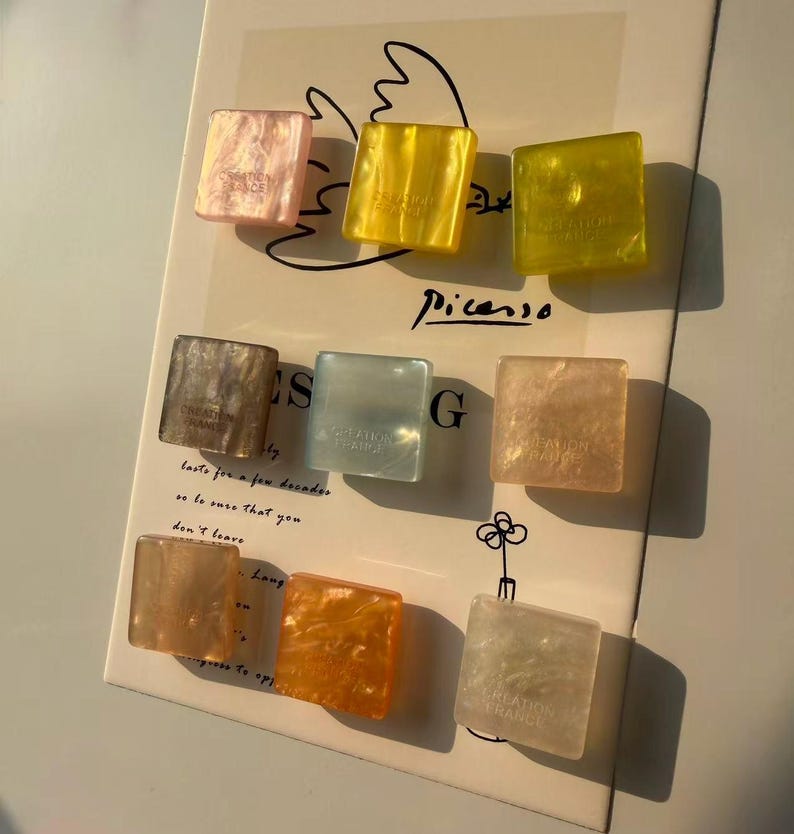 May include: Nine square-shaped hair clips in various colors, including pink, yellow, and orange. Each clip has a marbled effect and the text "CREATION FRANCE". The clips are arranged on a paper with the word "Picasso" and other text.