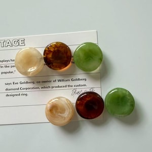 May include: Six circular, smooth, polished buttons arranged on a white card with printed text. The buttons feature marbled patterns in cream, dark brown, and translucent green. Each button has a glossy finish and a rounded, convex surface profile.