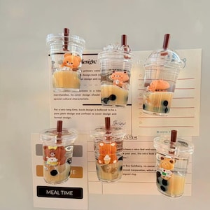 Puede incluir: Seis vasos de plástico transparente, cada uno con una pajita marrón, llenos de líquido amarillo y perlas de boba negras, que se asemejan al bubble tea. Cada vaso tiene una pequeña figura de oso. El texto "MEAL TIME" es visible.