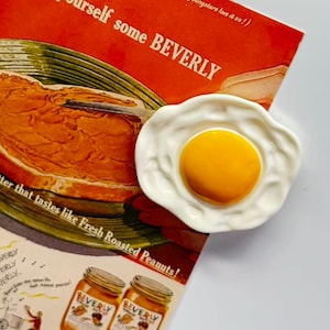 Może przedstawiać: Biały ceramiczny talerz w kształcie jajka z jasnożółtym żółtkiem. Talerz znajduje się obok starej reklamy masła orzechowego "BEVERLY", z słoikami i kanapką. Reklama zawiera tekst "spread yourself some BEVERLY".