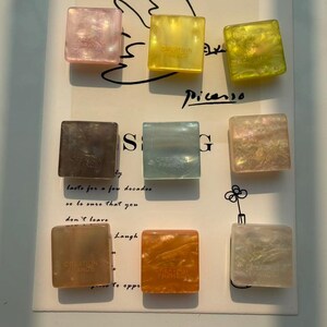 May include: Nine square-shaped decorative objects in various colors, including pink, yellow, green, brown, blue, and orange. Each object has a marbled or iridescent appearance and the text "CREATION FRANCE". The objects are arranged on a white surface with handwritten text.