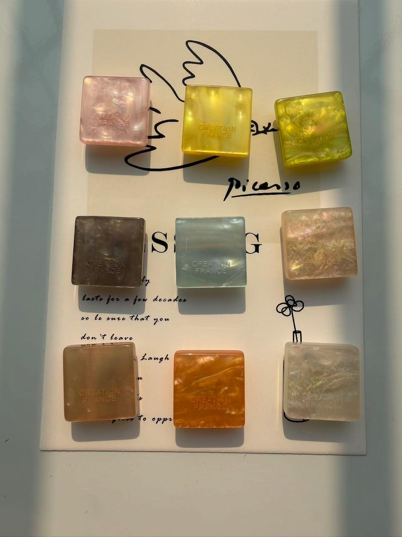 May include: Nine square-shaped decorative objects in various colors, including pink, yellow, green, brown, blue, and orange. Each object has a marbled or iridescent appearance and the text "CREATION FRANCE". The objects are arranged on a white surface with handwritten text.