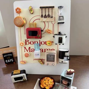 Puede incluir: Un tablero magnético blanco con el texto Bonjour Madame y varios imanes de electrodomésticos de cocina en miniatura, incluyendo una tostadora roja, una arrocera blanca, una licuadora, un dispensador de agua, una cafetera e imanes de comida.