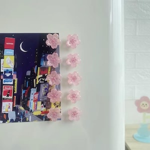 May include: Ten translucent light pink flower-shaped magnets arranged in two vertical columns. The magnets are securing a rectangular print depicting a nighttime cityscape with illuminated billboards and a crescent moon against a dark sky.