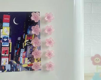 May include: Ten translucent light pink flower-shaped magnets arranged in two vertical columns. The magnets are securing a rectangular print depicting a nighttime cityscape with illuminated billboards and a crescent moon against a dark sky.