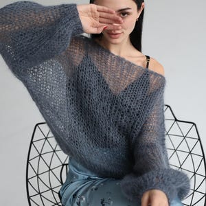 May include: A sheer, open-weave, blue-gray sweater with long sleeves. The sweater is worn over a dark top and paired with a blue floral-print skirt. The model is seated in a black wire chair.