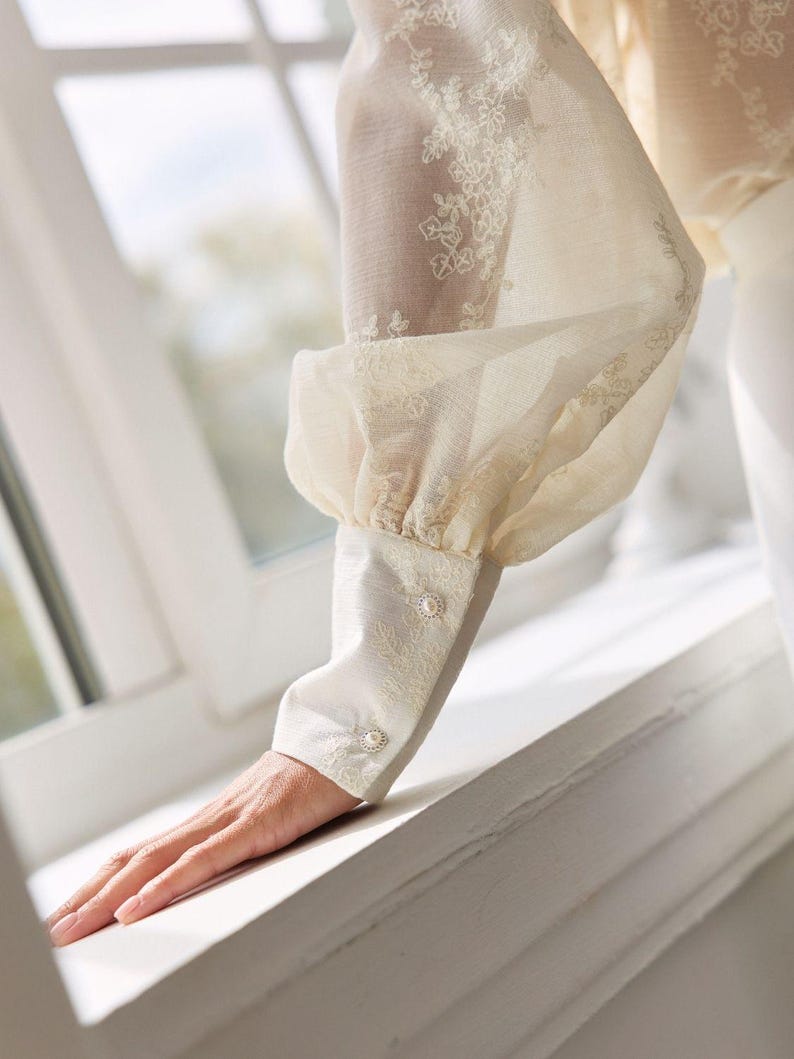 May include: A cream-colored long-sleeve blouse featuring sheer fabric with delicate white floral embroidery. The sleeve has a voluminous puff design gathered at the wrist into a fitted cuff, which is secured by two small, round, pearl-like buttons.