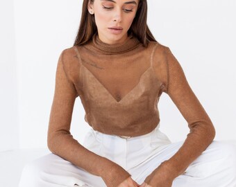 May include: A sheer brown long-sleeve turtleneck top with a visible V-neck camisole underneath, paired with white wide-leg trousers and tan pointed-toe heels. The top features a fine knit texture and a fitted silhouette with long sleeves.