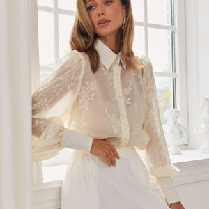May include: A sheer cream-colored long-sleeve blouse featuring delicate white floral embroidery, a structured collar, and buttoned cuffs. The garment is paired with solid white trousers, creating a clean and layered look with translucent fabric textures.