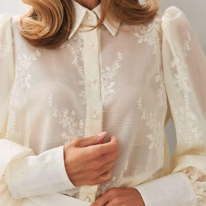 May include: A sheer cream-colored blouse featuring delicate white floral embroidery, long puff sleeves with wide cuffs, a classic collar, and a front button-down closure with small round buttons. The fabric is lightweight and semi-transparent.