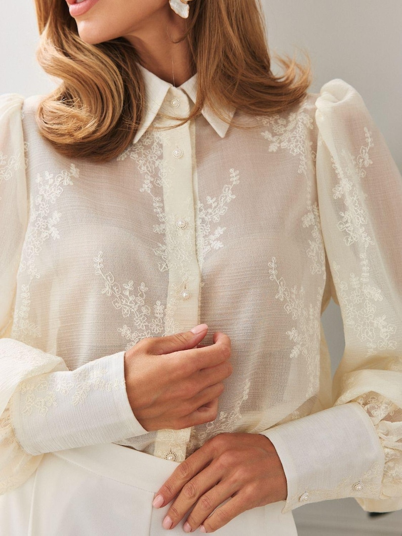 May include: A sheer cream-colored blouse featuring delicate white floral embroidery, long puff sleeves with wide cuffs, a classic collar, and a front button-down closure with small round buttons. The fabric is lightweight and semi-transparent.