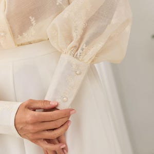 May include: A cream-colored long-sleeve blouse featuring sheer fabric with delicate white floral embroidery. The garment has voluminous sleeves, structured cuffs with two small pearl-like buttons, and is paired with a solid white skirt.