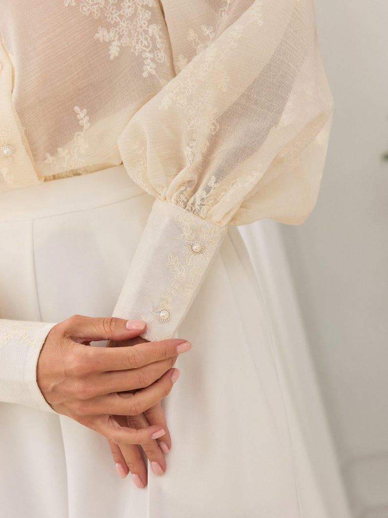 May include: A cream-colored long-sleeve blouse featuring sheer fabric with delicate white floral embroidery. The garment has voluminous sleeves, structured cuffs with two small pearl-like buttons, and is paired with a solid white skirt.