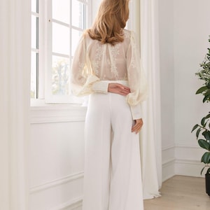 May include: A long-sleeved blouse featuring sheer fabric with white floral embroidery and voluminous sleeves, paired with solid white high-waisted wide-leg trousers. The blouse has buttoned cuffs and a high neckline, while the trousers fall straight.