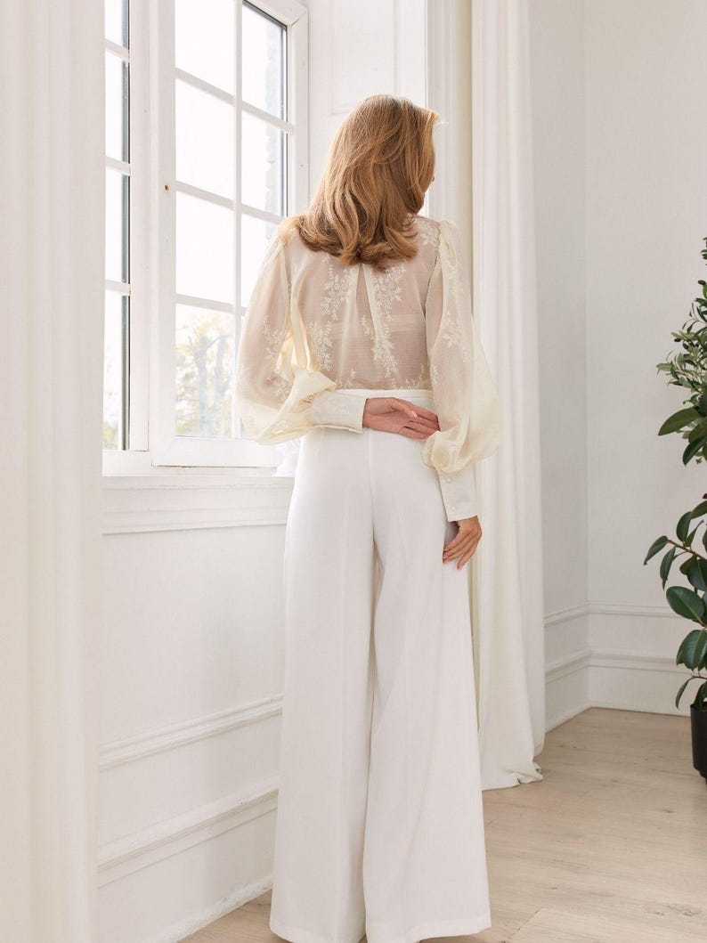 May include: A long-sleeved blouse featuring sheer fabric with white floral embroidery and voluminous sleeves, paired with solid white high-waisted wide-leg trousers. The blouse has buttoned cuffs and a high neckline, while the trousers fall straight.