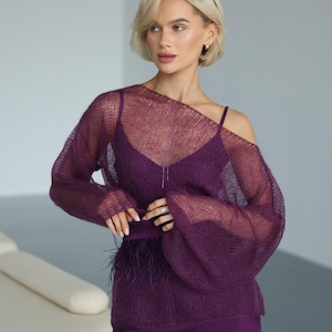 May include: A deep purple ensemble consisting of a sheer, loose-knit mohair sweater with wide sleeves worn over a matching camisole. The camisole features a straight neckline and a waistline accented with dark purple feathers, paired with a skirt.