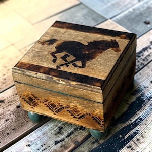 Può includere: Una scatola quadrata in legno con finitura marrone scuro, caratterizzata dalla silhouette di un cavallo al galoppo bruciata sul coperchio. Il pannello frontale presenta un motivo geometrico e la scatola poggia su quattro piccoli piedini arrotondati color ottanio.