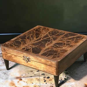 May include: A square wooden box with four tapered legs and a metal latch on the front. The top surface features a dark, intricate, branching fractal pattern burned into the wood, contrasting with the lighter brown grain of the box's sides.