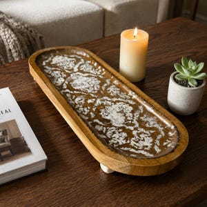 May include: An elongated oval wooden tray with a dark brown finish and a distressed white damask pattern on the interior surface. The tray is supported by four small, light-colored feet and features a smooth, polished rim around the edge.