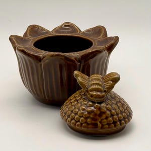 May include: A brown ceramic container with a flower-shaped body and a lid featuring a bee-shaped handle. The container has a dark interior and a textured exterior. The lid is off to the side.