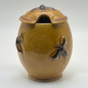 Puede incluir: Un tarro de miel de cerámica con tapa y una pequeña abertura para una cuchara. El tarro es de color marrón cálido con detalles de abeja marrón oscuro. La tapa tiene un asa en forma de abeja.