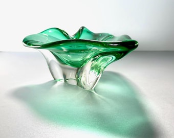May include: A clear glass bowl with a vibrant green hue. The bowl has a unique, organic shape with a ruffled rim and three curved legs. The glass is transparent, allowing light to pass through and create a shadow.