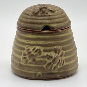 Op de afbeelding: Een keramische honingpot in de vorm van een bijenkorf. De pot is een gedempte olijfgroene kleur met horizontale ribbels en reliëf bijenontwerpen. De deksel heeft een kleine opening voor een lepel en een bijenontwerp.
