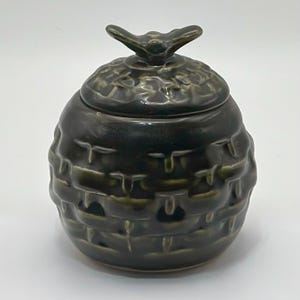 Może przedstawiać: Ciemnozielony ceramiczny słoik z pokrywką, przypominający ul. Słoik ma teksturowaną powierzchnię z wzorem tkanym. Pokrywka zwieńczona jest uchwytem w kształcie pszczoły. Słoik jest prawdopodobnie używany do przechowywania miodu.