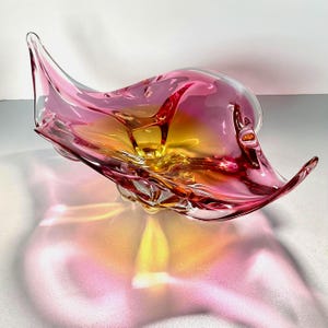 May include: A hand-blown glass bowl featuring an organic, asymmetrical shape with flowing, undulating edges. The glass transitions from a deep magenta pink at the outer rims to a vibrant, warm yellow hue at the centre, creating a translucent effect.