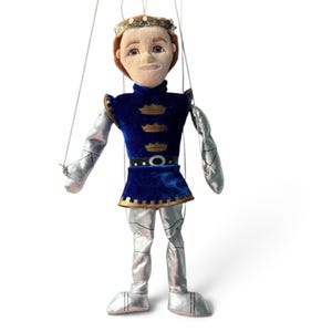 Puede incluir: Una marioneta de príncipe con cabeza de felpa, cabello castaño y una corona dorada. La figura viste una túnica de terciopelo azul con tres emblemas de corona dorados, un cinturón negro y mangas y pantalones de tela plateada metálica.