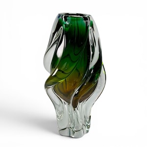 May include: A tall glass vase featuring a swirling, organic design with a clear base that transitions into deep emerald green and amber tones. The structure has a thick, heavy appearance with fluid, wave-like curves and a smooth, polished finish.