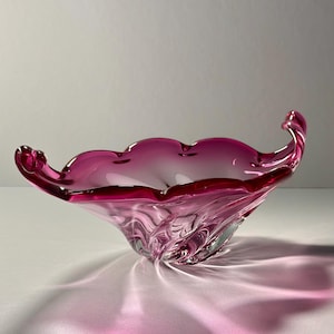 May include: A magenta-coloured, hand-blown glass bowl. The bowl has a scalloped rim and a swirling, textured design. The form is reminiscent of a flower, with elegant, curved handles. The bowl is made of glass.