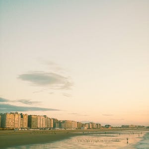 Może przedstawiać: Scena nadmorska z plażą, budynkami i ptakiem w locie. Niebo przechodzi od jasnoniebieskiego do ciepłego, złotego odcienia. Mokry piasek odbija niebo i budynki. Obraz ma estetykę vintage, przypominającą film.