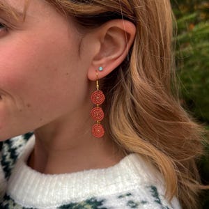 May include: Coral-coloured, three-tiered dangle earrings with a spiral design. The earrings are suspended from gold-coloured hooks. The earrings are a warm, earthy tone, and are shown on a person's ear.