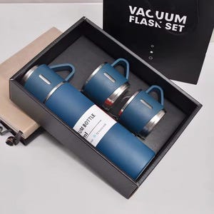 Peut inclure: Un ensemble de thermos bleu avec deux tasses assorties, présenté dans un coffret noir. Le thermos et les tasses ont des accents et des poignées argentés. L'ensemble comprend une étiquette qui indique "Vacuum Bottle". Un sac noir avec "Vacuum Flask Set" est en arrière-plan.
