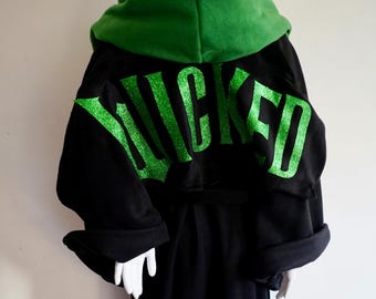 May include: Black robe with a green hood and the word "WICKED" in glittery green letters on the back. The robe has long sleeves and a belt. The robe is displayed on a white surface.