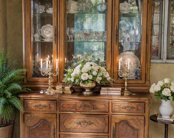 Könnte beinhalten: Ein großer, antiker Holzschrank mit Glasschränken und Schubladen. Der Schrank ist mit Glaswaren und Porzellan gefüllt. Ein Blumengesteck und Kandelaber schmücken die Oberseite. Ein Farn in einem Korb und ein kleiner Tisch mit Blumen befinden sich auf beiden Seiten.