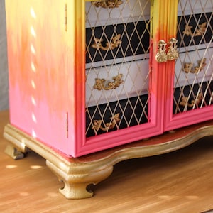 May include: A small, decorative cabinet with a gradient paint job in shades of yellow, orange, and pink. The cabinet has two doors with gold-colored hardware and a decorative base. The interior features white drawers and black backing.
