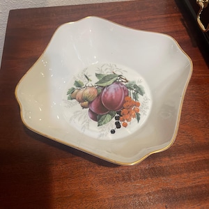 May include: A white porcelain serving dish with a gold rim and a square shape. The center of the dish features a colorful illustration of various fruits, including plums, berries, and other produce.