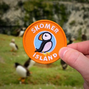 May include: Circular orange patch featuring a puffin bird illustration within a blue circle, with the text "SKOMER ISLAND". The puffin is depicted in black, white, pink, and yellow. The patch is held against a blurred background of green grass and puffins.