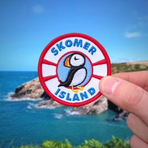 May include: A round embroidered patch featuring a puffin bird illustration. The patch has a red and white striped border with the words "SKOMER ISLAND" in blue lettering. The puffin is black, white, and orange, with yellow feet.