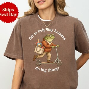 May include: Espresso-colored t-shirt featuring a frog riding a scooter with a bag of school supplies. The text reads "Off to help tiny humans do big things." A red circle with the text "Ships Next Day" is in the upper left corner.