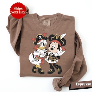 A brown t-shirt with a cartoon image of Minnie Mouse and Donald Duck dressed as pirates, along with a red "Ships Next Day" sticker.