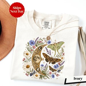 May include: An ivory t-shirt featuring a graphic print of a golden crescent moon decorated with lunar phases, surrounded by various wildflowers, lavender sprigs, and three moths in shades of green and brown, with small star accents throughout.