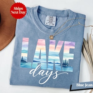 A blue t-shirt with the word "LAKES" printed on it, along with a necklace and a hat, all placed on a white surface.