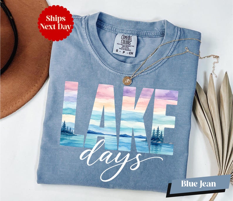 A blue t-shirt with the word "LAKES" printed on it, along with a necklace and a hat, all placed on a white surface.