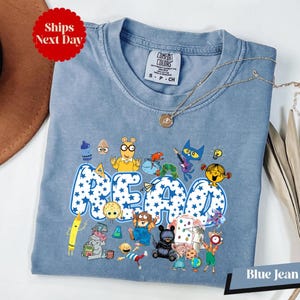 A blue t-shirt with a cartoon character design and the word "READ" printed on it, along with various other cartoon characters. The t-shirt is displayed on a white surface, and there is a brown hat and some decorative leaves in the background.